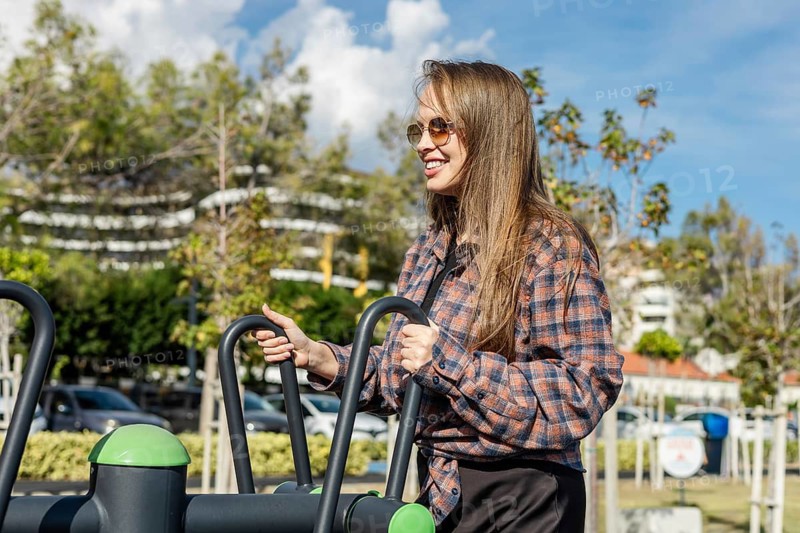 A young Caucasian woman with long brown hair exercises outdoors on fitness equipment. She wears sunglasses and a plaid shirt, surrounded by greenery and buildings