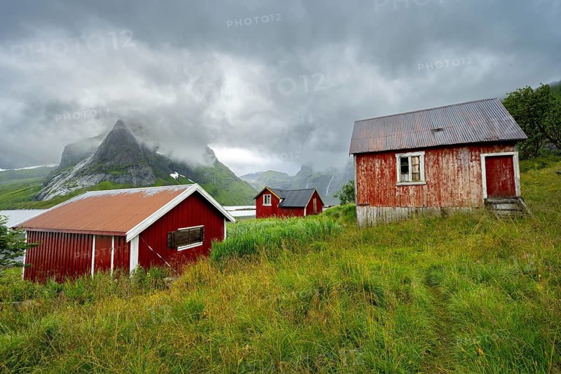 Red wooden houses in green mountain landscape with dramatic clouds, Vindstad, Reinefjord, Lofoten, Norway