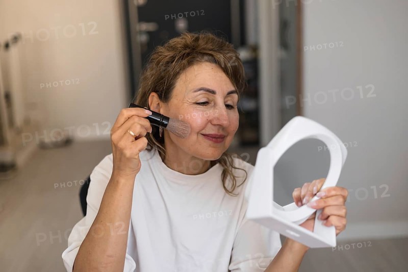 A middle aged woman sits comfortably in bright room at home, using brush to apply makeup on her cheek while gazing into handheld mirror. She appears focused and relaxed, showcasing her beauty routine