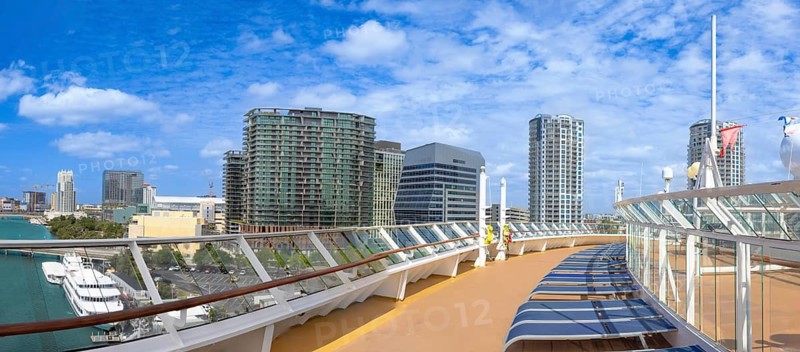 Panoramic view from a cruise ship deck in Tampa, Florida, USA. Sun loungers, glass railing, and modern downtown skyscrapers under a bright blue sky with clouds