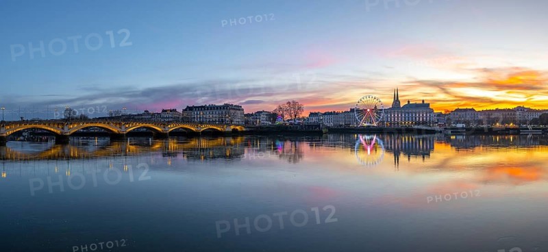 Bayonne skyline at christmas with an illuminated bridge and ferris wheel reflecting on the calm adour river under a colorful sunset twilight, festive urban riverside panorama