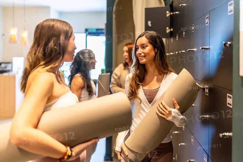 Two smiling women are holding rolled yoga mats and talking in a modern locker room at a wellness studio, preparing for a group fitness class and healthy lifestyle