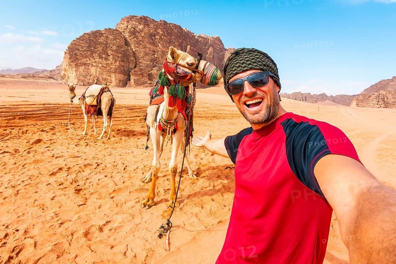 Smiling man traveler taking selfie standing near saddled camel orange sand valley sandstone rock mountains background summer vacation adventure tourism Wadi Rum Jordan