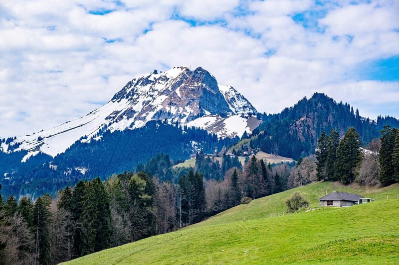 Snowy mountain above green meadow with hut surrounded by trees and cloudy sky, Villars-sous-Mont, Canton of Fribourg, Switzerland
