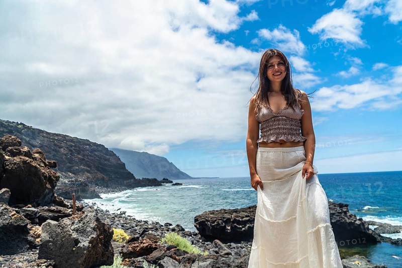 Young woman smiling while standing on the rugged volcanic coastline of el hierro, spain, enjoying scenic ocean views during her summer travel vacation