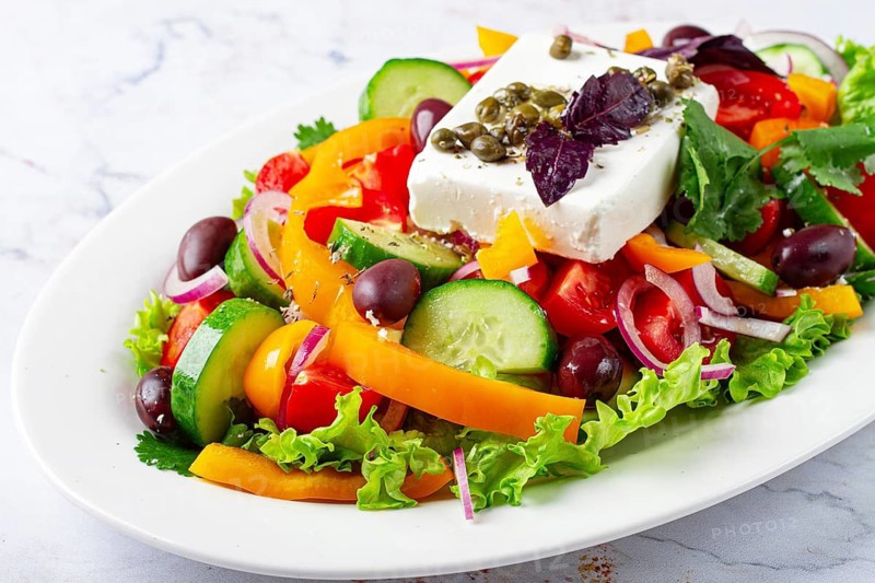 This dish features a salad made with a mix of lettuce, peppers, cucumbers, red onion, and olives. It is topped with feta cheese and capers