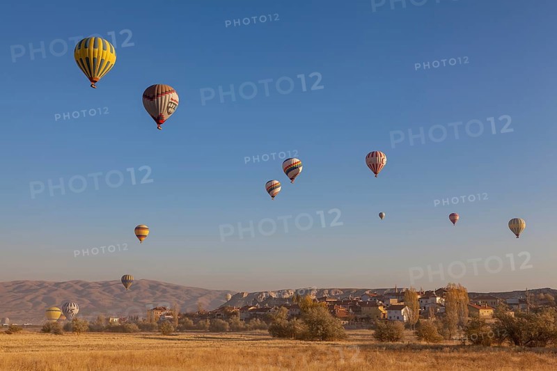Several hot air balloons soar into the bright blue morning sky over an idyllic village and vast countryside, Cappadocia, Turkey