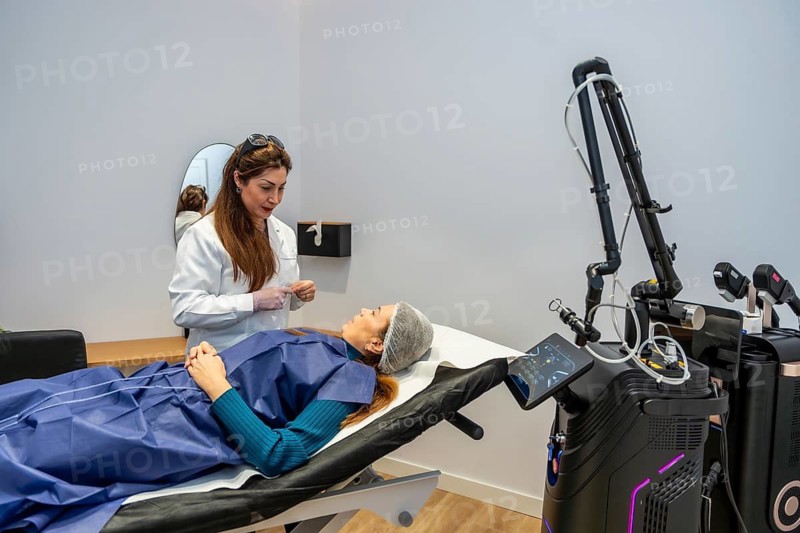 Doctor in white coat and gloves consulting female patient in disposable gown and hairnet, lying on procedure bed beside advanced laser equipment in modern aesthetic clinic