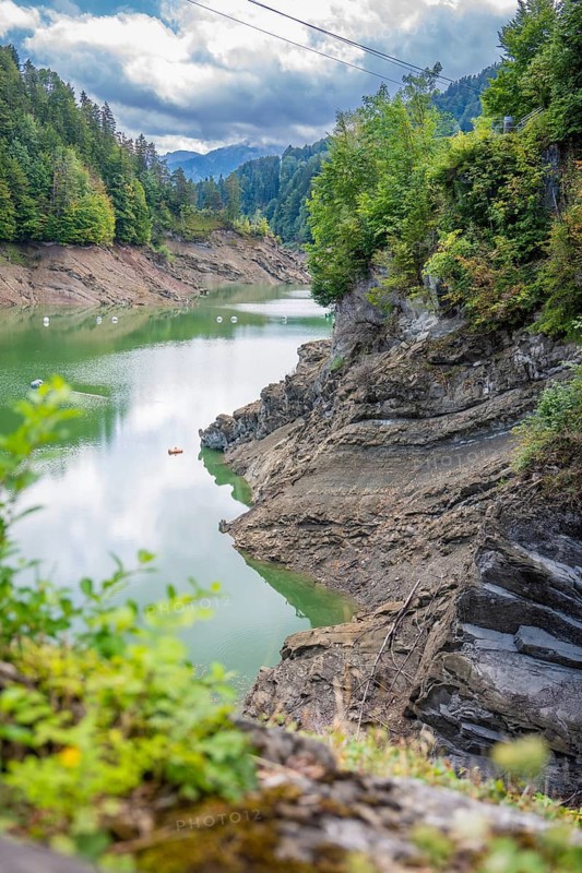 Clear lake with rocky coast, surrounded by green trees and calm water surface, Krumbach, Vorarlberg, Austria