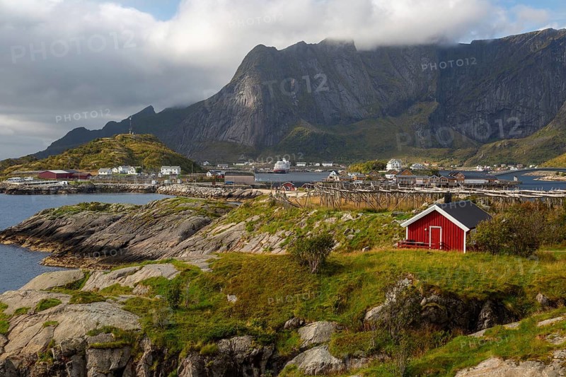 Lonely red Rorbuer hut on a green meadow with a view of pure and dried fish racks in front of cloudy mountains, Reine, Lofoten, Norway