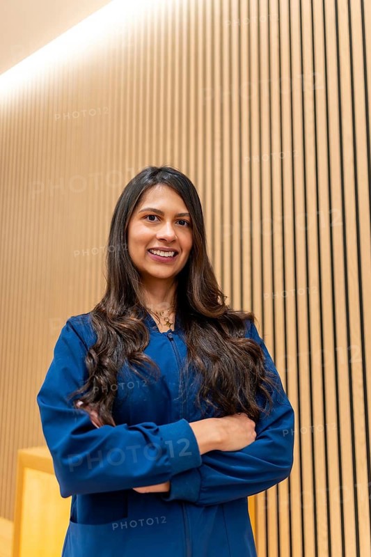 Hispanic professional woman wearing blue scrubs, confidently smiling with arms crossed, standing against a modern vertically slatted wooden wall in an aesthetic clinic or medical spa setting