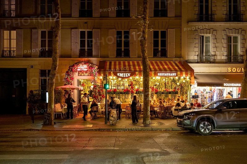 Nocturnal, summer street scene in front of a traditional French bistro, outdoor dining, with guests at outdoor tables, atmospheric lighting with lanterns and fairy lights, and a distinctive striped awning in front of a historic house façade, Paris, Paris department, Île-de-France region, France