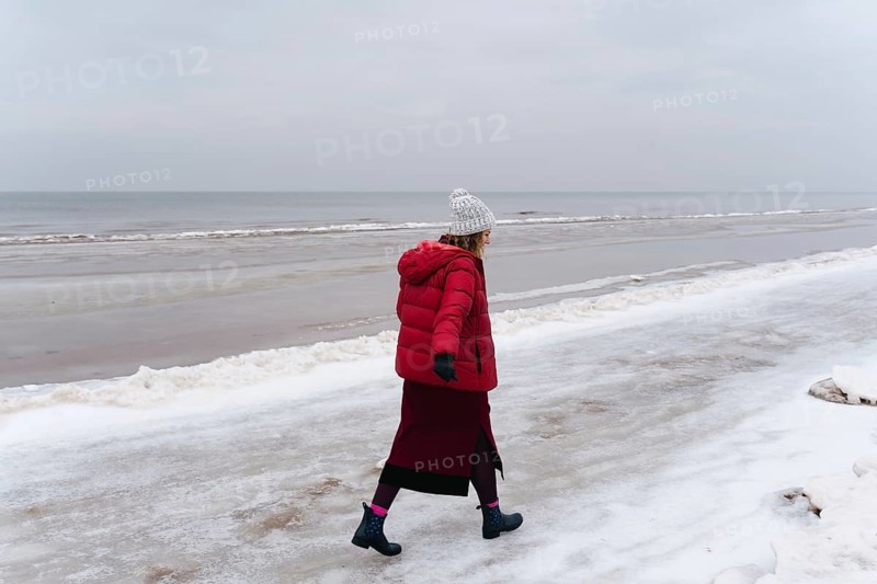 A woman in warm clothes walks along the sea in winter, A woman in a red coat walks alone on a snowy beach by the ocean under a cloudy sky, Jurmala, Latvia