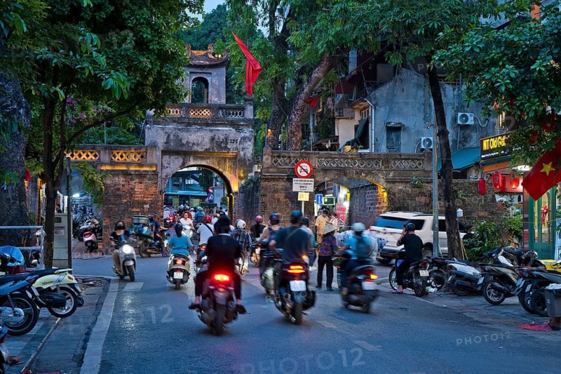 City gate during the evening rush hour on a busy street with mopeds, scooters, motorbikes and passers-by, surrounded by historic buildings and trees, Historic City Gate in the Old Quarter of Hanoi Vietnam