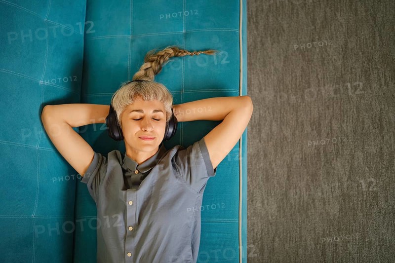Woman lies on blue couch with headphones on, eyes closed, and hands resting behind head. Soft light fills room, creating comfortable atmosphere for relaxation and enjoyment of music