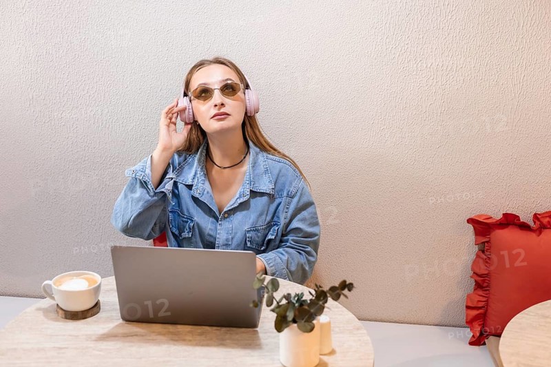 Young Caucasian woman with long brown hair wearing pink headphones, sitting at a table with a laptop and a cup of coffee, in a cozy indoor setting