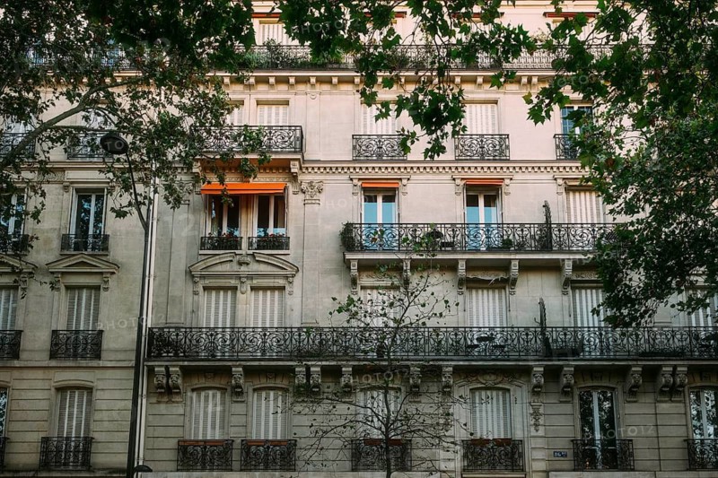 Classic Parisian residential building façade from the Belle Époque with ornate balcony grids and window awnings in front of a historic row of houses in the city center, urban street scene with trees in the foreground. Paris, Paris Department, Île-de-France Region, France