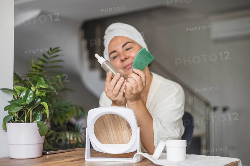 A young Hispanic woman with a towel on her head holds a gua sha tool and a serum bottle. A mirror and a plant are on the table, creating a serene beauty setting