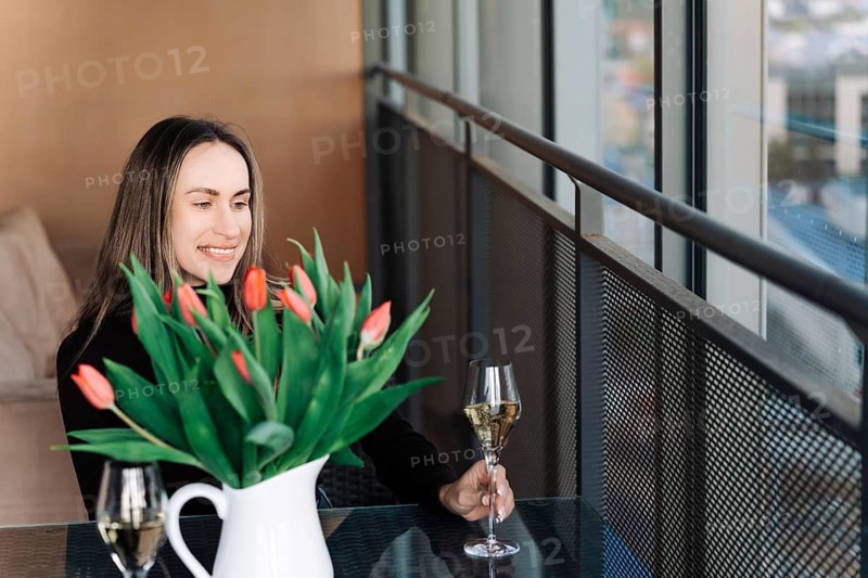 A young woman sits at a table by the window and holds a glass of champagne. There is a vase of flowers on the table, A woman in a black sweater smiles, holding a glass of wine near a window with tulips in a vase