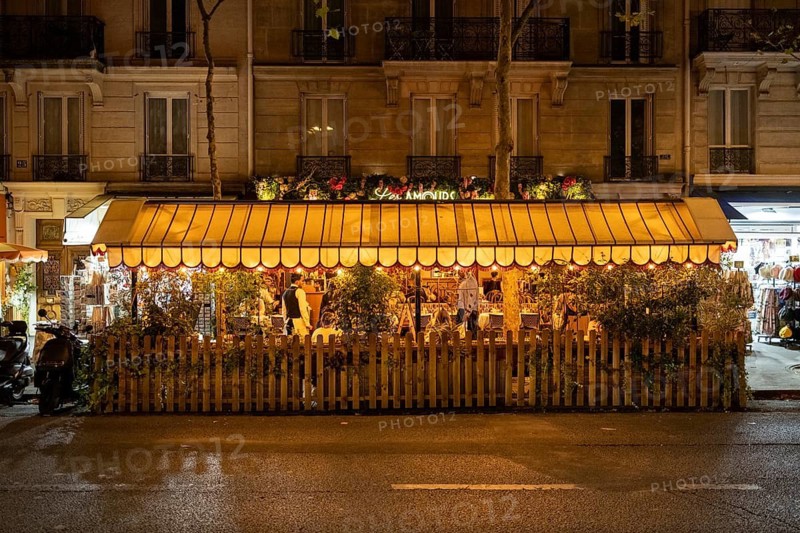 Nocturnal, summer street scene in front of a traditional French bistro, outdoor dining, with guests at outdoor tables, atmospheric lighting with lanterns and fairy lights, and an awning in front of a historic house façade, Paris, Île-de-France region, France