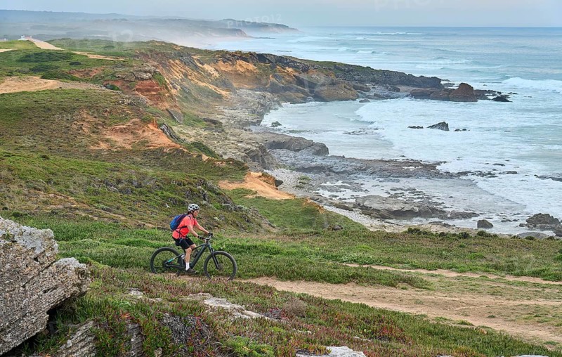Active woman riding her electric mountain bike at the rocky and sandy coastline of the atlantic ocean in Porto Covo, Portugal