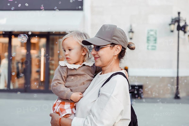 A woman holds her young daughter close as they watch bubbles float in the air. The city square is bustling with a warm autumn atmosphere, creating a joyful scene of bonding
