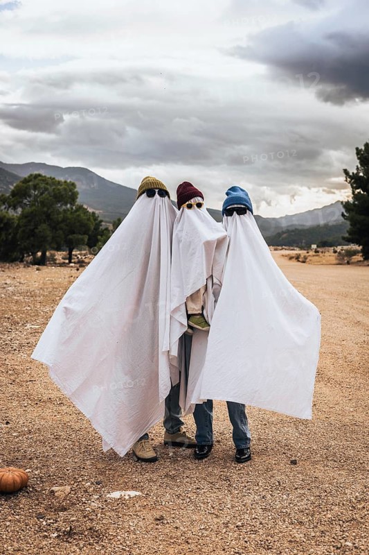 Three children dressed as ghosts for Halloween, wearing white sheets and hats. They stand together in a rural area with mountains in the background