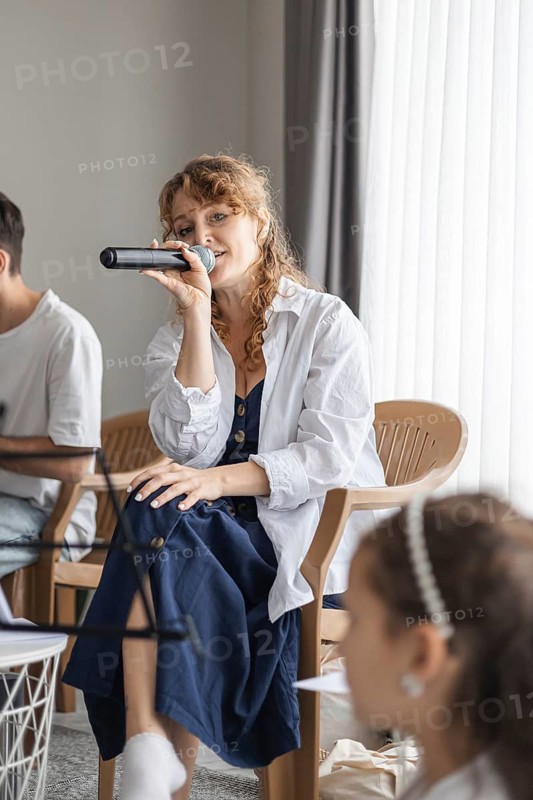A young Caucasian woman with curly hair sings into a microphone at a home concert. Two children listen attentively, creating a warm family atmosphere