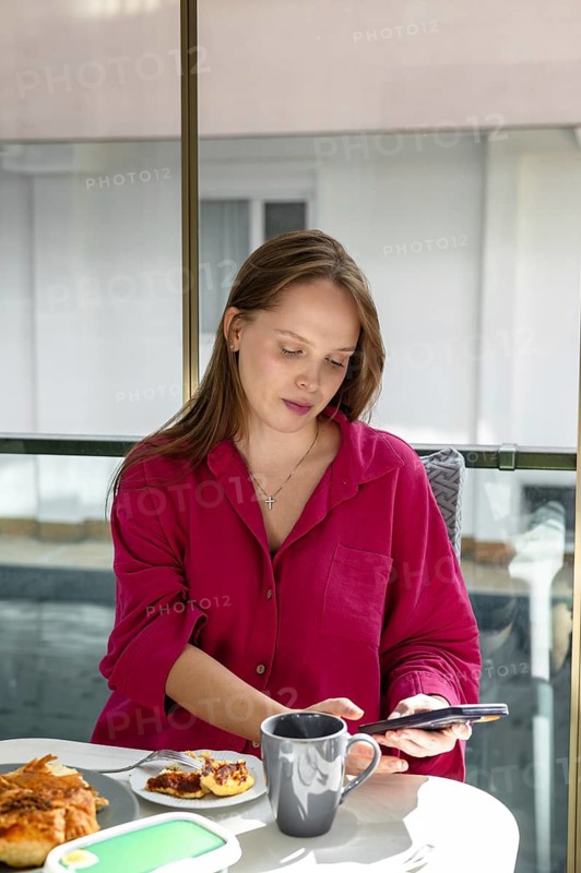 Woman sits at a table, looking at her phone. She has a cup next to her and some food on a plate. The setting is bright and indoors. She wears a pink shirt