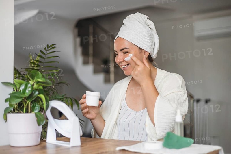 Woman applying skincare while enjoying a beverage in a cozy indoor setting in the morning