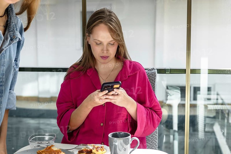 Woman sits at a table, looking at her phone. She has a cup next to her and some food on a plate. The setting is bright and indoors. She wears a pink shirt