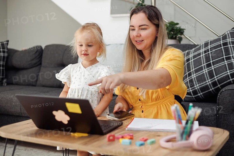Young Caucasian woman with blond hair and a small blond girl using a laptop on a table in a living room, home education concept
