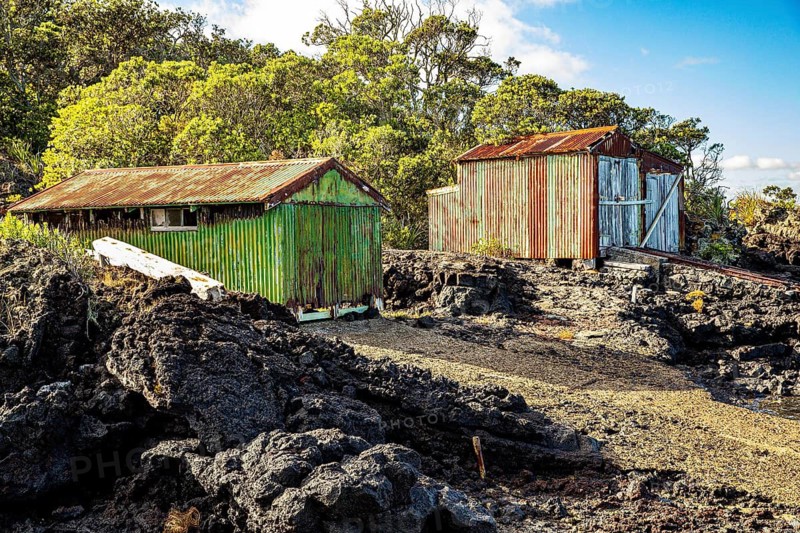 Two corrugated iron boatsheds, one green and one rust red, sit among black lava rock on the shore of Rangitoto Island, weathered remnants of the historic bach settlements that once served the island's holidaymakers, New Zealand
