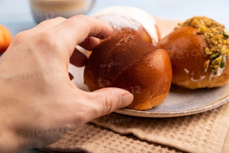 Maritozzo traditional Italian bun dessert with hand, cup of coffee, on blue wooden background with orange linen textile, close up, side view, selective focus