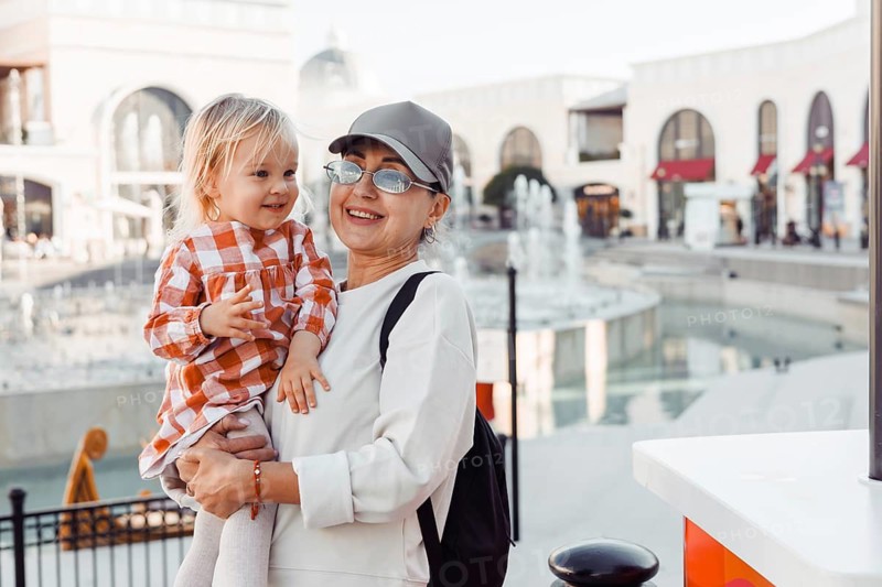 A grandmother smiles as she holds her young granddaughter close by a city fountain. The pair enjoys a sunny afternoon, surrounded by shops and a vibrant atmosphere
