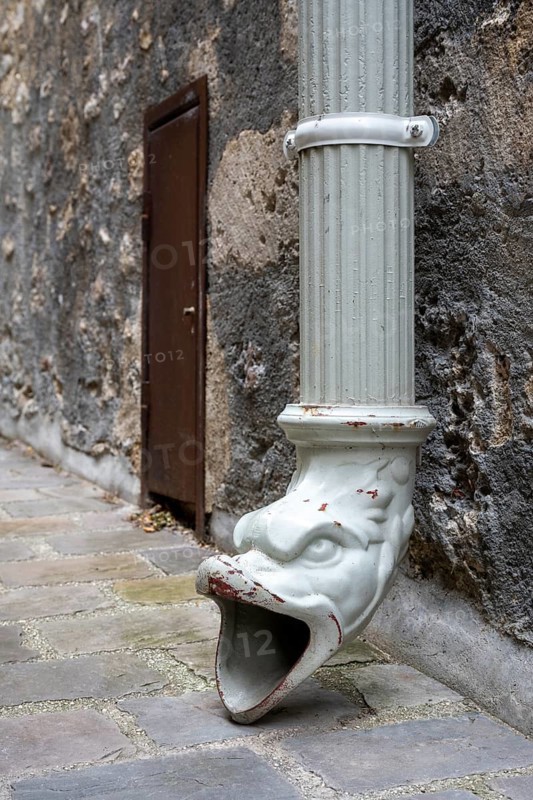 Detailed view of an antique rainpipe outlet in the shape of a decorative gargoyle figure. The decorated metal pipe directs water onto the paved floor next to a house entrance. Troyes, Aube Department, Grand Est Region, France