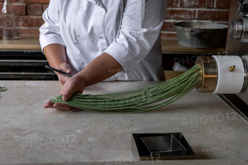 Process of woman preparing handmade pasta. Woman's hands working with dough to make pasta. Chef making green dough blanks to make craft pasta. Chef in white jacket preparing food in restaurant kitchen. Small business in the food industry with a focus on high quality and healthy food concept. A woman chops pasta in a special food processor for noodles, green dough with the addition of spinach