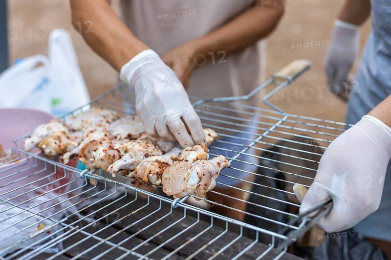 Hands placing marinated chicken pieces on a grill. The scene shows a close-up of food preparation for a barbecue. Gloves are worn for hygiene