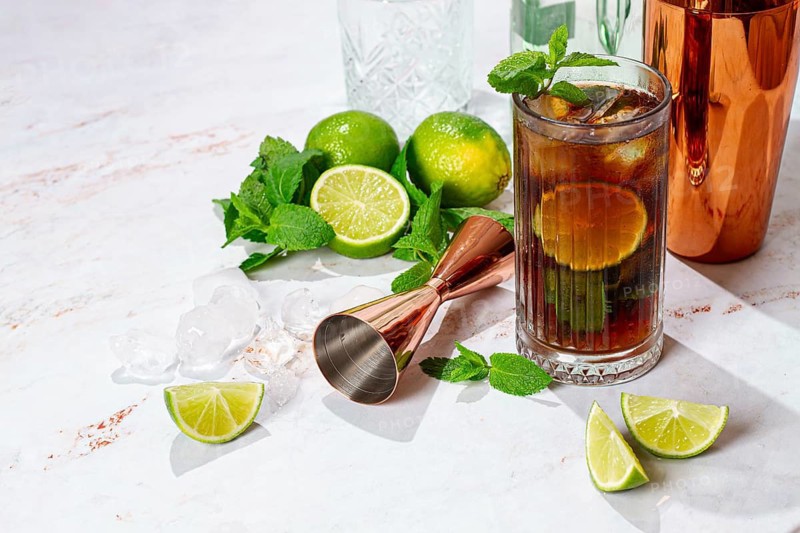 A tall glass filled with a dark soda drink shows slices of lime and fresh mint on a white table. A copper shaker sits in the background with other glassware nearby