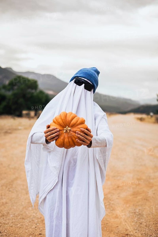 Person dressed as a ghost holding a pumpkin outdoors during Halloween celebration. Homemade costume, autumn traditions and playful seasonal atmosphere.