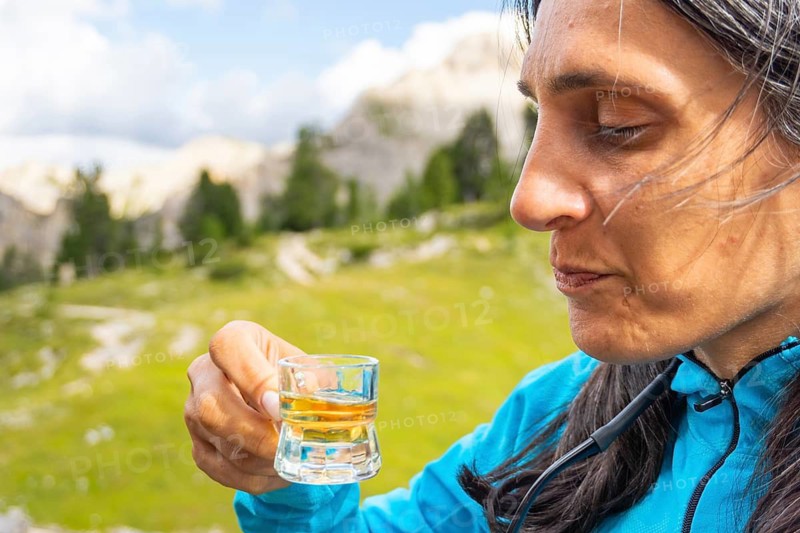 Close up woman traveler drinking local herbal spirit schnapps or grappa shot glass wooden terrace green mountain valley backdrop Dolomites Italy. Fragrant, grape-based pomace home made brandy