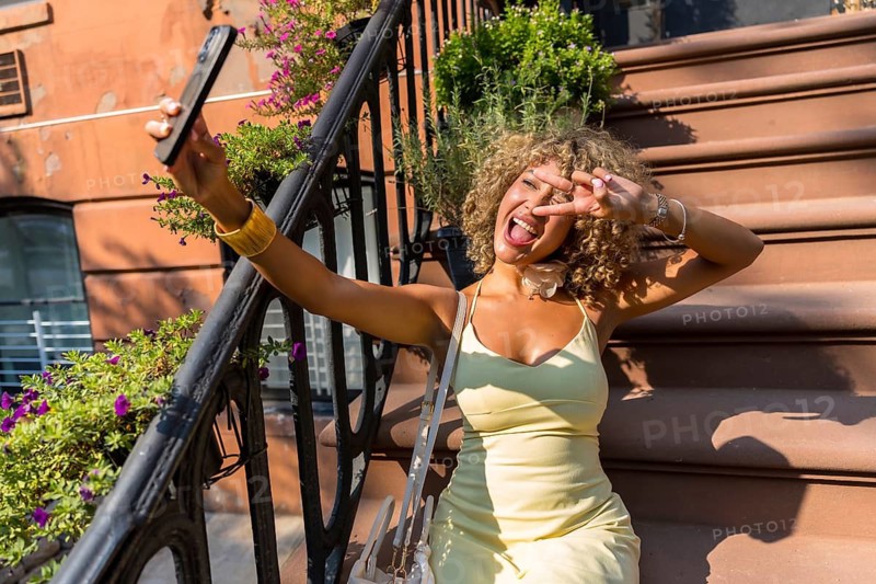 Young woman with curly hair expressing joy and making a peace sign while taking a selfie on a brownstone stoop in a manhattan neighborhood during summer