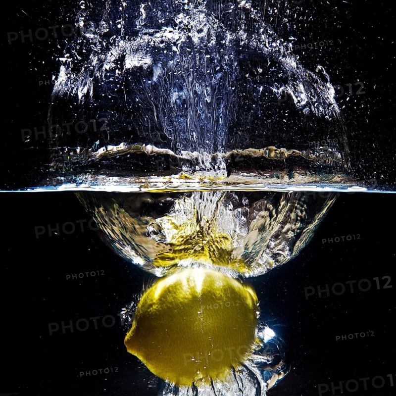 A fresh yellow lemon (Citrus limonum) falls into the water, creating a splash and air bubbles, against a black background
