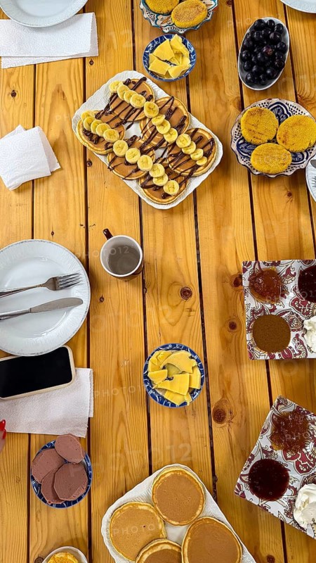 Overhead view showing a spread of pancakes with banana and chocolate syrup. Fresh fruit. Cheese. Jam. And various pastries arranged on a rustic wooden table. Creating a vibrant scene of a morning meal