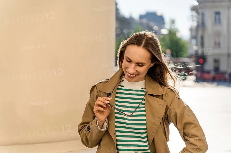 A woman smiles and holds sunglasses while walking in the city, A smiling woman in a trench coat holds sunglasses in an urban setting during a sunny morning, Vilnius, Lithuania, A woman smiles while holding sunglasses outdoors in a city wearing a trench coat and striped shirt, A smiling woman holding sunglasses in an urban setting, dressed casually in a trench coat and striped shirt, A woman smiling in a trench coat outdoors in a city setting under sunlight, Woman in a trench coat and striped shirt holds sunglasses while smiling on a sunny city street, Vilnius, Lithuania