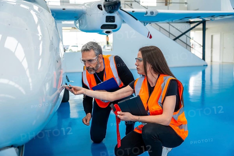 Two aviation technicians wearing safety vests and goggles inspecting the fuselage of a light aircraft in a hangar, carefully checking for details as part of routine maintenance and safety procedures