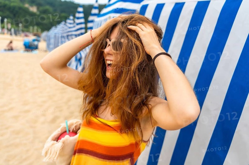 Young reddish haired woman laughing heartily. Playfully running hands through her hair. Wearing stylish sunglasses and a striped top