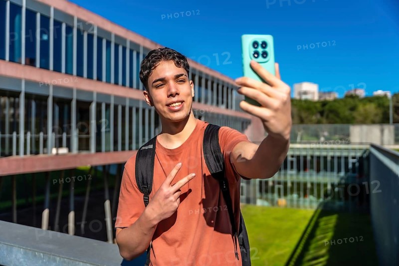 Young male student smiling while capturing a selfie with his smartphone, showing a peace sign on a modern university campus with a clear blue sky, embracing youth, technology, and education
