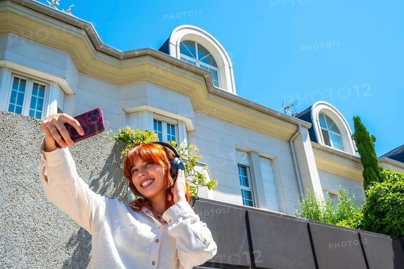 Young red haired woman wearing headphones smiles while taking a smartphone selfie in front of a modern urban building on a sunny day, enjoying music and social sharing