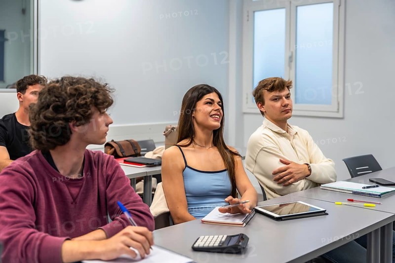 Happy students of diverse ethnicities participating in a university lecture, learning on their desks with notebooks, tablets, and pens in a bright, modern classroom